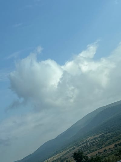 This media file depicts a landscape scene, seemingly in Mirovo, Bulgaria. The image is oriented vertically, with the top of the frame showing a clear blue sky transitioning into a large, soft cloud formation. The cloud is predominantly white with some gray undertones, suggesting it may be moderately dense.

The lower portion of the image reveals a sloping hillside or mountain. The terrain is covered in dense vegetation, appearing as a mosaic of green and darker shades, indicating a forested area. There are also visible patches that seem to be cleared land, possibly for agriculture or pasture, with some lighter brown or yellow areas. A faint, indistinct line running diagonally across the hillside could be a road or path.

The time of day appears to be daytime, given the bright sky. The presence of clouds and the general lighting suggest possibly overcast or partly cloudy weather, though the sky in the upper part of the frame is clear. There are no people or specific objects clearly identifiable in the scene. The overall mood is serene and natural, focusing on the expansive sky and the textured landscape. No visible text is present in the image.