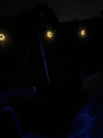 This nocturnal image, captured in Villa Pasquali, Italy, depicts an outdoor scene characterized by profound darkness. In the upper middle section, a dark-colored telescope is mounted on a tripod with blue-tinted legs, positioned against a slightly lighter, overcast night sky with faint silhouetted vegetation. Three identical circular decorative lights are affixed to an unseen dark vertical surface, likely a fence or wall, across the middle ground. Each light emits a warm yellow glow, casting intricate radial patterns resembling a mandala or starburst. In the lower left foreground, a human hand, primarily obscured by shadow, holds a lit cigarette, evidenced by its distinct glowing red ember. To the right in the lower foreground, parts of a person's legs, covered in dark blue fabric (possibly jeans), are visible, suggesting a reclining or seated posture. The overall environment is dark, indicating a late-night setting, possibly in a private garden or backyard, with stargazing or celestial observation as a likely activity.