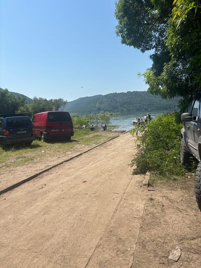 A dirt road runs alongside a body of water, bordered by trees and a hillside. Two vans and an SUV are parked on the grassy shoulder of the road, with a boat visible in the water. The sky is clear and blue.