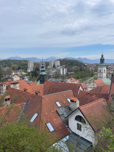 The image presents an elevated perspective of Skofja Loka, Slovenia, characterized by red-tiled rooftops and buildings. The backdrop includes a clear sky with scattered clouds and distant mountains with traces of snow. Visible architectural features include church towers with green spires. Modern residential buildings are present amidst the historical structures. Green foliage suggests springtime. There are no visible people in the image.