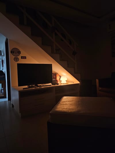 The image captures an interior scene, likely at night, within a residence or hospitality establishment in Cabo Frio, Brazil. The primary focus is a warmly lit corner next to a staircase, creating a cozy atmosphere.

No people are visible in the scene. On the left, a staircase ascends upwards into the darkness, with its underside revealing wooden or metal supports decorated with circular designs.

The central feature is a brightly illuminated niche or corner, where a distinctive white lamp with a curved shade emits a warm, orange glow. Another similar decorative lamp or object stands beside it. Below these lamps, a large, dark rectangular object, possibly a TV screen or a framed item, is mounted on the wall. Above this, a shelf holds a box prominently labeled "CHIVAS 12" and another box that resembles a Nintendo Switch package, along with other small items.

The wall space adjacent to the dark rectangular object is decorated with several circular stickers or badges. Visible text on these includes "RUTA 40 HISTÓRICA," "5194," "VINHOS DO ALGARVE," "Bariloche," and "MANTEQUERA," suggesting travel, regional pride, and potentially connections to Argentina and Portugal.

To the right of the illuminated corner, wooden cabinets or built-in storage units are visible, extending vertically. The upper and lower portions of the image are dimly lit, showing dark walls and ceilings, and hinting at an adjoining room or hallway at the bottom.

The dim overall lighting, contrasted with the bright, warm artificial light from the lamp, indicates that it is nighttime or late evening. The ambiance is tranquil and inviting, with no specific activity taking place beyond the static display of an interior space.