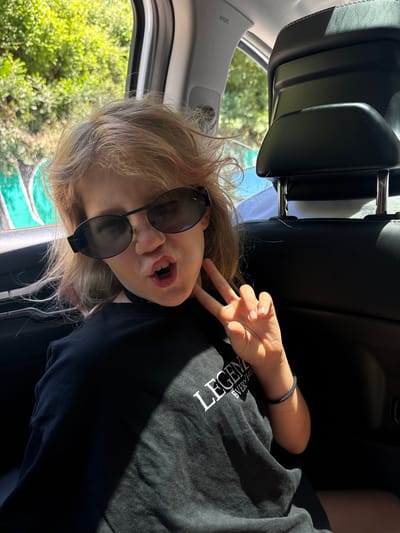 A child is in the back seat of a car, making a peace sign with their hand. They are wearing sunglasses and a black shirt with white text on it. The car appears to be moving, as greenery is visible outside the window. The location is unclear, and there's no information to suggest it's in Kruče - Kërruçi, Montenegro.