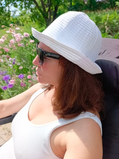 A woman wearing a white hat and sunglasses sits in a chair outdoors, facing left. She has brown hair and is wearing a white tank top. Behind her is a garden with pink and purple flowers. The scene is illuminated by sunlight. The location is Vyazovka, Russia.