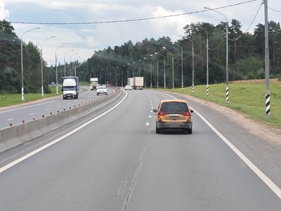The image depicts a multi-lane highway in Mednoe, Russia, under an overcast sky. The road is paved with asphalt and marked with white dashed lines. A concrete barrier separates the lanes, with red reflectors visible along its top edge. Numerous streetlights with curved poles line the roadside, interspersed with striped black and white marker posts. Dense green forest borders the right side of the highway, while the left side shows a grassy verge. Several vehicles are present on the highway, including a white truck with a blue cargo container, two white cars, a white truck with a flatbed trailer, and a yellow car in the foreground. The yellow car, a hatchback, is moving away from the viewer and displays a license plate that appears to be from Russia. The overall scene suggests a typical highway environment in a forested area.
