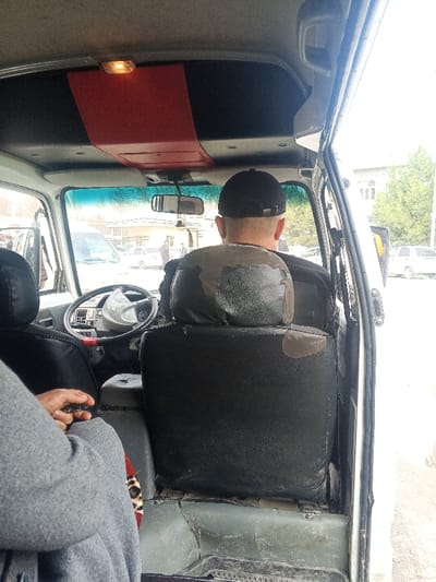 The image depicts an interior view from a passenger seat within a white commercial van or minibus, looking forward towards the front and out the windshield. The location is specified as Laish, Uzbekistan.
**People:** Two individuals are present. In the immediate foreground on the left, the lower torso and left arm of a person are visible, wearing a gray long-sleeved garment. Their right hand holds a small, dark, rectangular object. A subtle leopard print pattern is visible near their lap. The second individual, presumably the driver, is seen from behind, wearing a black baseball cap and a dark top. Their hair is short on the back of the head.
**Objects and Vehicle Interior:** The vehicle's interior features a light-colored ceiling with a prominent central red panel, which houses a single illuminated rectangular light fixture. A rearview mirror hangs centrally from the ceiling. The front windshield has a blue tint strip at the top. The steering wheel is light-colored with a red accent on its lower rim. Both visible seats are covered in dark, worn material; the driver's headrest and upper backrest exhibit significant degradation with patches of original material exposed. The floor is covered with worn, dark gray-green matting. The front passenger-side door is open or ajar, revealing its white exterior frame.
**Exterior Environment:** Through the windshield and open door, a daylight street scene is visible. Several vehicles are present, including a white van/minibus partially seen on the left and a white sedan in the middle distance. A light-colored building with multiple windows and green trees are visible in the background, suggesting an urban or suburban setting.