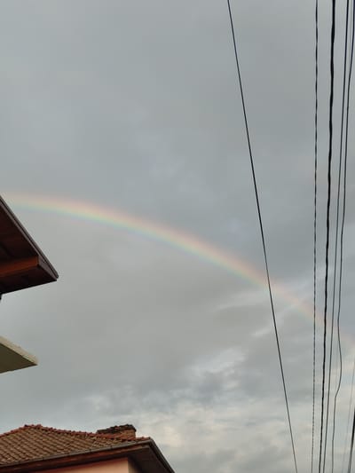 The image depicts a cloudy sky with a rainbow arcing across it. Several power lines stretch horizontally across the upper portion of the frame. In the foreground, the rooftops of two houses are visible, one with terracotta tiles and the other a darker material. The scene appears to be set in a residential area in Pletena, Bulgaria, likely during the daytime given the presence of the rainbow, suggesting recent rain and sunlight. The overall mood is peaceful and serene, highlighting a natural phenomenon against an urban backdrop. There is no visible text in the image.