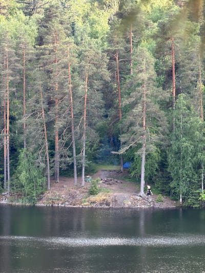 The image depicts a serene natural landscape with a dense forest bordering a dark, reflective body of water. The forest consists of tall, slender pine trees with a mix of deciduous trees and undergrowth. On the right side of the frame, a clearing in the trees reveals a campsite with a small blue tent, a campfire pit with embers, and a few scattered items. Two individuals are visible near the campsite; one is sitting on the ground, and the other, a child, is being carried on someone's shoulders. Further to the left, closer to the water's edge, a person is seated on the bank. The water itself is calm, with ripples creating a shimmering effect on its surface. The overall scene conveys a sense of peaceful recreation in a wilderness setting. The location is identified as being in the city of Ruch'i, Russia.