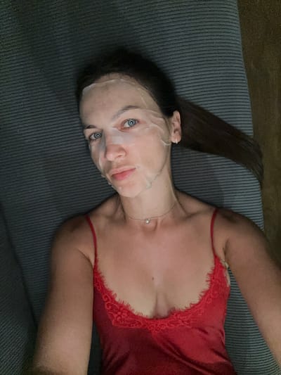 A woman is lying on a ribbed, dark grey couch, with her head tilted to the left. She is wearing a red silk slip with lace trim. A sheet mask covers her face, leaving her bright blue eyes and pink lips visible. Her long, dark hair is spread out on the couch to her left. A thin silver necklace with a small diamond pendant is visible around her neck. A ring is on her left hand, which is resting near her head. The lighting suggests it is indoors, possibly at night or in a dimly lit room. The overall mood appears relaxed and self-care oriented. There is no visible text in the image.