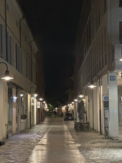 The image depicts a narrow street in Ravenna, Italy, at night. The street is paved with cobblestones that reflect the light from street lamps lining both sides. The buildings on either side are multi-story structures with shopfronts at ground level. On the left, there are windows with closed shutters and awnings. On the right, the building features a more modern facade with columns and a pixelated image of a Space Invader on the wall. In the middle of the street, a dark-colored car is parked. Further down the street, a person is walking away from the camera. There are also several tables and chairs set up outside a building, some of which are covered with umbrellas. The overall atmosphere is quiet and dimly lit, suggesting a late-night scene.