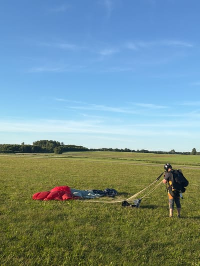 This is a photograph taken outdoors, likely in Limbaži, Latvia, on a clear day with a bright blue sky and scattered wispy clouds. The sun is shining, indicating it is daytime, possibly late afternoon due to the warm lighting on the grass. The scene is a grassy field with a line of trees in the distance, suggesting a rural or park-like setting.

The main subject is a person, dressed in a black and yellow skydiving suit, helmet, and goggles, crouched down on the grass. The person appears to be a skydiver, preparing to pack or having just unpacked their parachute. The parachute itself is visible on the ground, partially spread out and colored red, white, and blue. The parachute lines are visible, connecting to the skydiver's harness. The skydiver's body language suggests focus and concentration on the task at hand. The image has a vertical orientation, with the sky occupying the left side and the ground, skydiver, and parachute on the right. No text is visible in the image.