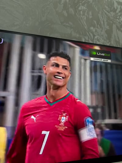 The image shows a close-up of a television screen displaying a soccer player wearing a red jersey with the number 7. The player has dark hair and is smiling. The jersey has a crest on the chest and a Puma logo on the left side. There is also a white armband on the player's right arm with blue text and graphics. In the upper right corner of the screen, there is a "LiveBall" logo with Russian text below it that says "ПРЯМОЙ ЭФИР" (meaning "Live Broadcast"). The background of the screen appears to be a stadium with blurred figures of other people. The image is taken from an angle, and the television screen is displaying a broadcast. There is no information in the image to suggest it is located in Osh City, Kyrgyzstan.