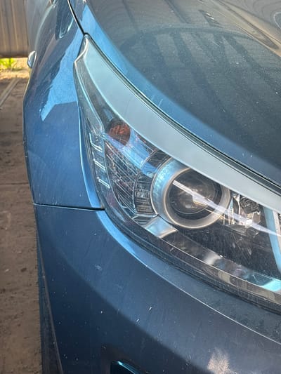 This media file is a close-up, low-angle shot of the front right headlight and fender of a dark blue car. The car appears to be dusty, with visible specks of dirt and some minor scratches on the surface, particularly on the fender. The headlight itself is complex, featuring a projector lens with metallic accents and a clear, ribbed lens cover. An amber turn signal is visible within the headlight assembly. The car is positioned outdoors, as suggested by the dappled sunlight reflecting off the metallic paint and the presence of what appears to be vegetation and a weathered wooden fence in the background, indicating a rural or semi-rural setting. The lighting suggests it is daytime, possibly a sunny day due to the bright reflections. No people are visible in the frame. The overall impression is of a vehicle that has been used and exposed to the elements. The metadata indicates the location is Sovkhoza "Razdol'e", Russia. There is no visible text in the image.