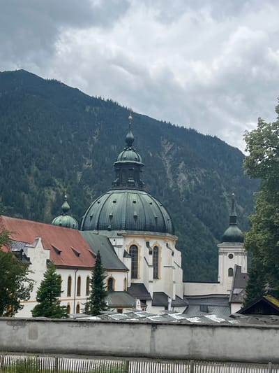 The image captures a portion of the Ettal Abbey complex in Ettal, Germany, set against a backdrop of a lush, forested mountain under a cloudy sky. The architecture of the abbey is prominently featured, with its distinctive green-domed structures and red-tiled roofs. A large central dome, characteristic of Baroque architecture, dominates the mid-ground, while a smaller dome and a bell tower are also visible. The building is painted in white with yellow accents around the windows and cornices, showcasing its ornate design. Evergreen trees are interspersed around the abbey, adding to the natural setting. In the foreground, a low, grey wall runs horizontally across the frame, with a wooden fence situated in front of it, partially obscured by the wall. The overall scene depicts a historic religious complex nestled within a mountainous landscape.