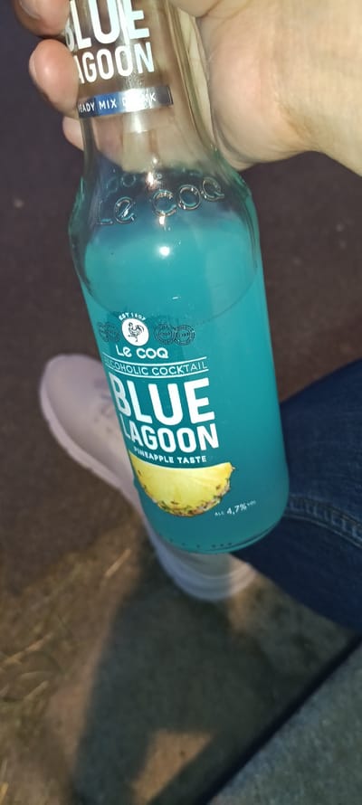 A close-up, angled shot shows a hand holding a clear glass bottle filled with a bright blue liquid, identified as "BLUE LAGOON" alcoholic cocktail. The bottle has white text labels indicating "READY MIX DRINK," "Le Coq," "ALCOHOLIC COCKTAIL," and "PINEAPPLE TASTE." A sliced pineapple is depicted on the label. The text "ALC 4,7% vol" is also visible. The hand holding the bottle appears to be an adult's, with visible skin and fingernails. The bottle is positioned over a white sneaker and dark blue denim fabric, suggesting the person is seated or standing. The background is out of focus and appears to be a dark, outdoor environment, possibly a street or park at night, with some visible ground texture. There is no specific indication of the city of Linz, Austria in the image.