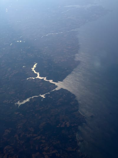 This is an aerial photograph of the coastline near Fouesnant, France, with a body of water on the right side of the image and land on the left. The land appears to be a mix of agricultural fields and forested areas, with some settlements visible. The coastline is irregular, with bays and inlets. The water is a deep blue, and there appears to be some haze or mist over the water near the coastline. The sun is shining, causing reflections on the water and highlighting certain areas of the land. The overall impression is one of a rural, coastal landscape.