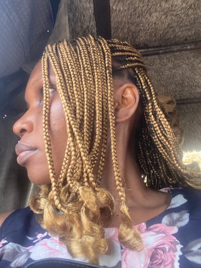 A person with braided blond hair lies down, strands covering the eyes. The braids are neat near the scalp but curl at the ends. The person's ear is visible, and their skin tone is dark. A floral-patterned garment appears on the right. A blurry background suggests an indoor setting.