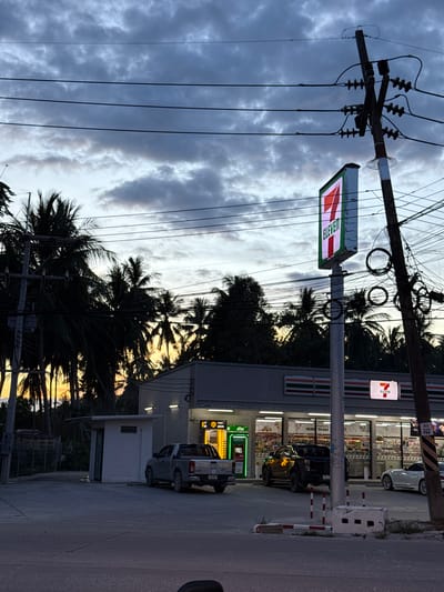 The image depicts a 7-Eleven convenience store at dusk in Baan Nai Suan, Thailand. In the foreground, power lines are strung across a utility pole. Two 7-Eleven signs are visible, one perpendicular to the road and another attached to the store building. Palm trees obscure some of the background. Several vehicles are parked near the storefront.