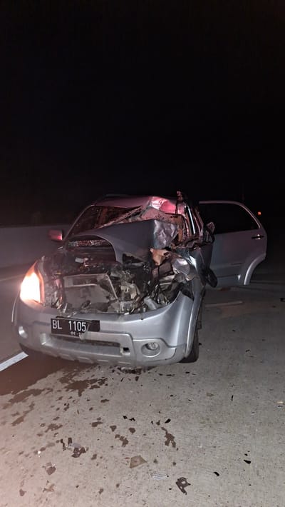 A silver car has been in a severe accident, with its front end completely crushed. The car is lying on its side on a concrete surface, possibly a road or a roadside. Debris from the accident is scattered around the vehicle. The car's hood is open, revealing further damage to the engine compartment. A license plate is visible on the front of the car, reading "BL 1105" and "06-27". The scene appears to be illuminated by the car's headlights and possibly by artificial light sources, suggesting it is nighttime or very dark. The background is dark, indicating it is likely night. The concrete surface is stained with what appears to be liquids and small fragments, typical of an accident scene. The overall impression is one of destruction and chaos resulting from a significant collision. The location is Baksukon, Indonesia.