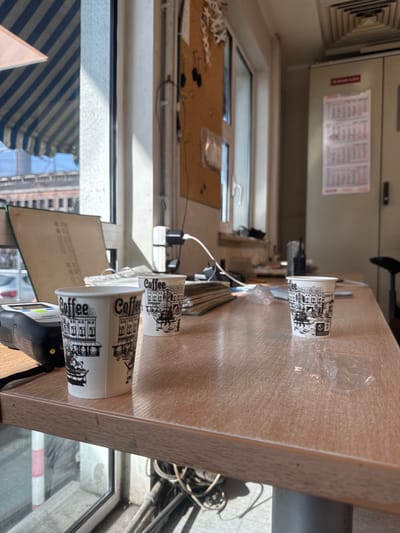 The image captures an interior scene, likely an office or service area, located in Obruchishte, Bulgaria. The vantage point is oblique, overlooking a light-colored, wood-grain table surface. Three white paper cups, each bearing black "Coffee" text and a cityscape illustration, are positioned on the table. To the left of the nearest cup, a black and grey electronic device with a green illuminated display element is visible. A crumpled, clear plastic wrapper rests on the table's right side.
Natural light streams into the space from a large window on the left. Through this window, a blue and white striped awning is seen, followed by a brick-colored building under a clear blue sky, with street-level elements like vehicles and poles partially observable. An open, light-colored book or document stands propped near the window. A wall-mounted corkboard, displaying various pinned items including keys and small white cutouts, is situated to the right of the window. Below the corkboard, multiple power cables and adapters are arranged.
In the background, a large, grey metal cabinet or locker stands against the far right wall. Two white calendar sheets and a red rectangular sign are affixed to its front. A square, metallic air vent is present on the ceiling in the upper right corner. No individuals are present in the scene.