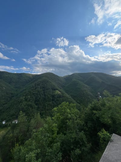 The image depicts a natural landscape dominated by verdant, tree-covered mountains under a partly cloudy blue sky. The foreground features the edge of a concrete structure, possibly a road or walkway, with some green foliage in the lower right corner. In the mid-ground, the slopes are densely wooded, with a few scattered structures, appearing to be houses with white roofs, visible nestled within the greenery on the left side. Further into the distance, the undulating mountain range stretches across the frame, showcasing its lush, forested contours. The sky above is a vibrant blue, interspersed with scattered white and gray clouds, some of which are illuminated by the sun, suggesting it is daytime. The overall impression is of a peaceful, rural mountainous region.