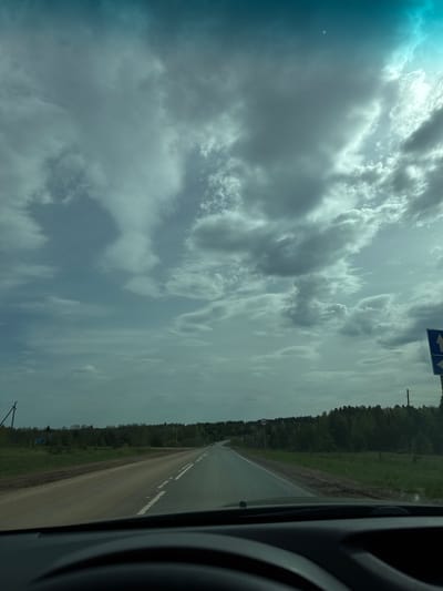 The image shows a view from inside a vehicle, likely a car, looking out onto a road. The road has a white line dividing the lanes and is flanked by green vegetation and a forest. The sky is filled with clouds. The location may be near Kamennyi Kliuch, Russia based on the prompt.