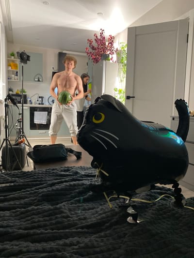 A shirtless man with curly blonde hair stands in a kitchen holding a watermelon. He is wearing light-colored sweatpants. Behind him, a woman in a white top and dark pants holds a green object, possibly another watermelon. A black cat-shaped balloon with yellow eyes and white whiskers is in the foreground, obscuring part of the scene. The kitchen appears to be in a residential apartment, with a stovetop, cabinets, and shelves visible. There are also various pieces of photography equipment, including tripods and lights, suggesting a photoshoot is taking place. The overall setting is indoors, in what appears to be a well-lit apartment kitchen.