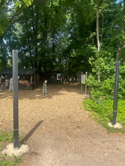 The image depicts an outdoor scene with a path covered in wood chips. On the left side of the path, there is a small market stall with green roofing and various items displayed. Several people are visible near the stall, including two children, one wearing a green hoodie and a white cap, and an adult.

Further into the scene, the path continues, leading towards more structures and dense green foliage. Several signs are posted on wooden posts on the right side of the path. The environment is heavily wooded with tall trees and lush undergrowth, suggesting a natural or park-like setting. The lighting indicates it is daytime, with shadows cast on the ground.

The provided information states the location is the city of Shlisselburg, Russia. While the image shows a market-like area within a wooded environment, there are no specific landmarks or details visible that definitively place it in Shlisselburg. It appears to be a casual outdoor market or event, possibly a recreational area.