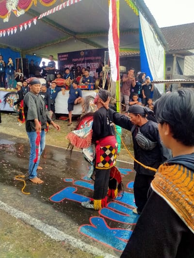 The image depicts an outdoor cultural performance in Tamansari, Indonesia, under a large, temporary canopy. The ground is a wet asphalt surface with scattered red petals and blue letters outlined in red painted on it, partially spelling "POTRO."
In the mid-ground, a central figure performs in a Jaranan (horse dance) costume, comprising a stylized, painted horse head worn at the waist, a red and black checkered sarong over black trousers, and red and yellow fringed leg adornments. This performer, wearing a black long-sleeved shirt, is being supported by a male figure to their right. The supporting figure wears a black long-sleeved shirt, black head covering, and a black fanny pack, with a yellow rope tied around his waist; his left hand is on the head of the horse dancer. To the left of the horse dancer, a barefoot male, wearing a black long-sleeved shirt and blue jeans with red markings, holds a red object, possibly a whip or ribbon, and faces slightly left. Another horse costume is partially visible behind him.
In the immediate foreground, the back of a person wearing a black hooded shirt with an orange and black patterned design on the shoulder is visible on the right.
In the background, under the canopy, an elevated stage hosts multiple individuals, predominantly young males in dark blue t-shirts with yellow logos, performing with traditional musical instruments including drums and gongs. A banner overhead reads "PAJAKAN JARANAN POTRO POTRO." Spectators, including adults and children, observe from around the stage, with some on the right holding mobile phones to record the event. Behind the performance area, a building structure is visible, partially obscured by a blue and white tarp. A large red and white flag (Indonesia's flag) on a pole stands prominently to the right of the stage.