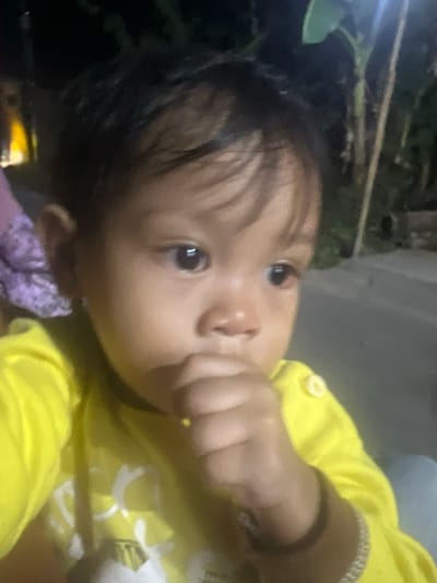 A close-up, low-light photograph shows a young child with dark hair and brown eyes wearing a bright yellow long-sleeved shirt. The child's right hand is near their mouth, with their fingers slightly raised. The background is dark and appears to be outdoors, with some blurred foliage and a light source on the left. There is also a suggestion of a paved surface in the background. The image quality is grainy due to the low light conditions. The child is positioned slightly off-center to the right, with their gaze directed towards the viewer's left. A thin, sparkling bracelet is visible on the child's left wrist. The setting is indoors or outdoors at night in the city of Wedarijaksa, Indonesia, indicated by the dark, unlit environment and visible vegetation.