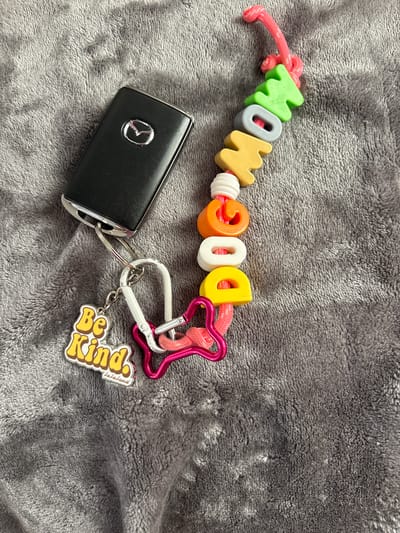 The image depicts a black Mazda car key fob attached to a keychain lying on a gray textured surface, possibly a blanket or soft fabric. The keychain features colorful letter beads spelling "MOM DOG" strung on a pink cord, interspersed with white spacer beads. A pink heart-shaped carabiner clip and a white carabiner are attached to the keychain, along with a "Be Kind" charm. The scene is not identifiable as being in Seal Beach, United States of America.