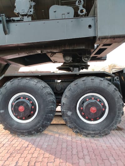 This is a close-up, low-angle shot of the massive tires and undercarriage of a large, dark-colored vehicle, likely a military truck or heavy equipment, possibly a mobile launcher. The vehicle is parked on a red interlocking brick paver surface. The tires are black with a rugged tread pattern. The wheel hubs are black with red bolt caps and a central red cap, surrounded by a white rim.
The undercarriage of the vehicle is complex, with visible mechanical components and structural beams. The overall impression is one of immense size and robust engineering. The lighting suggests it is either early morning or late afternoon, with the warm glow of the sun casting shadows and highlighting the textures of the bricks and tires. The sky is visible in the background, appearing bright and possibly clear.
There is visible text on the tires: "500-610 18.00-24".
The setting appears to be an outdoor, paved area. The presence of what looks like distant buildings or structures in the background, along with the overall industrial aesthetic of the vehicle, suggests a functional or industrial location. Given the provided location context of Baikonur, Kazakhstan, this could be a site related to space exploration or military operations, consistent with the heavy machinery depicted. No people are visible in the image. The mood is stoic and industrial, emphasizing the power and scale of the machinery.