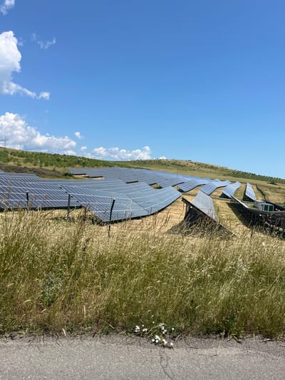 A solar farm is located on a hillside under a clear blue sky with scattered white clouds. The solar panels are arranged in multiple rows, covering the grassy slope. Some panels are angled upwards, while others appear to be flatter. In the foreground, tall dry grass lines a paved surface, possibly a road or pathway. The hillside is dotted with scattered green bushes and trees. A small green building or shed is visible among some of the solar panel arrays. The image captures a landscape view of renewable energy infrastructure in an outdoor, rural setting. The location is identified as the solar farm in Bryastovets, Bulgaria.