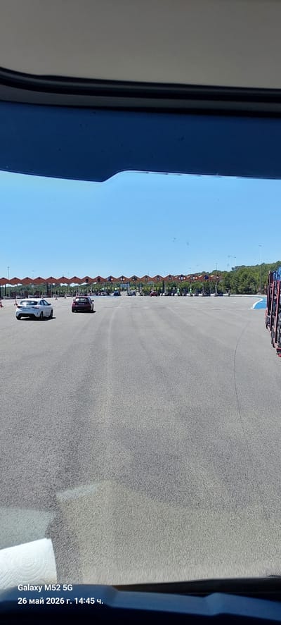 The image shows an outdoor scene, seemingly captured from inside a vehicle, likely in Cubelles, Spain. The view is of a toll road with multiple toll booths positioned in a line on the left side. Two cars are visible in the lanes leading to the booths: a white car closer to the camera and a burgundy car further away. The sky is clear blue. Along the left side beyond the toll booths, there's a line of trees. The image has some artifacts and is not perfectly clear, and there are parts of the vehicle interior visible on the left and right edges of the photo. The bottom right corner of the image has the phone's timestamp indicating that the picture was taken on May 26, 2026 at 14:45.