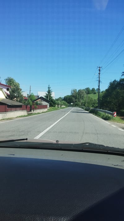 The image depicts a road in Deliatyn, Ukraine, from the perspective of someone inside a vehicle. The road is paved and has a white line marking the lane. On the right side of the road, there is a dark, textured surface, likely the dashboard of the vehicle. On the left side of the road, there are houses with red roofs and stone foundations, surrounded by greenery and trees. Power lines run along the left side of the road, supported by wooden poles. The sky is a clear, bright blue, indicating a sunny day. There is a sign on a curved pole near the side of the road with red and white stripes, possibly a warning sign. The overall scene suggests a rural or suburban setting during daytime. No people are visible in the image, and no text is discernable.
