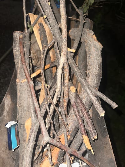 A pile of firewood, primarily consisting of dry branches and thicker logs, is arranged outdoors at night. The wood varies in color from light tan to dark brown and grey, with some pieces showing rough bark. Interspersed with the wood are thin, dry twigs. In the lower left corner, a partially visible blue and white cardboard box, likely containing cigarettes, is placed against a larger log. The background is dark, suggesting it is nighttime, and appears to be a natural or semi-natural outdoor setting. There is no indication of a specific location in the city of Samarkand, Uzbekistan.