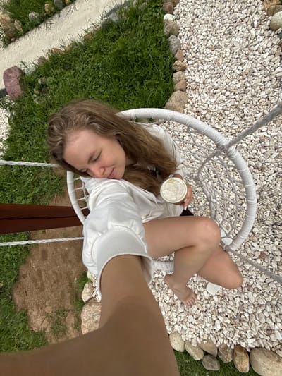 A young woman with long, wavy brown hair is reclining in a white rope hammock chair, holding a clear cup of a light-colored beverage. She is wearing a white hooded sweatshirt and white shorts. Her eyes are closed, and she appears relaxed, possibly napping or enjoying a quiet moment. The chair is suspended by thick white ropes.

The background consists of a mix of small white pebbles and larger, rounded stones. To the left and bottom of the image, there is lush green grass and some patches of dirt. The lighting suggests it is daytime, possibly overcast or late afternoon, as there are no harsh shadows. The setting appears to be an outdoor area, perhaps a garden or a yard, in Ryzhkovo, Russia. The overall mood is peaceful and serene.