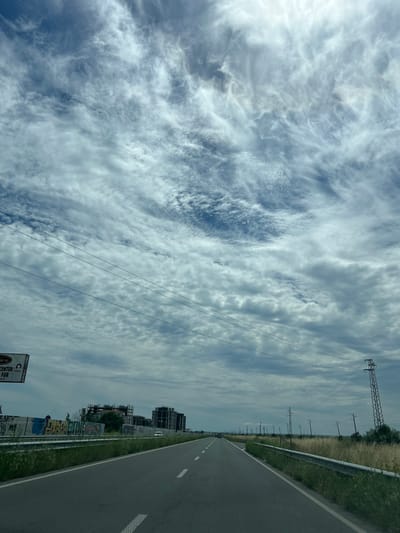 The media file depicts a roadside view under a cloudy sky, oriented vertically. The scene looks out over a paved road with white lane markings, suggesting a highway or major road. To the right of the road, there is a grassy verge and a metal guardrail. Beyond the guardrail, a field of tall, dry grass stretches towards a line of power poles and electrical wires that extend into the distance. In the background, partially obscured by the power lines, are several buildings, some appearing to be under construction with visible scaffolding. Graffiti is visible on a low wall or barrier closer to the foreground. The sky is filled with a dynamic pattern of white and gray clouds, with some patches of blue visible. The sun is not directly visible, but the brightness of the clouds suggests it is daytime, likely mid-morning or afternoon. The overall atmosphere is one of travel and passing through a semi-urban or developing area. There is a sign visible in the lower right corner with some text, but it is partially cut off and illegible. Another sign is visible near the bottom center, also partially obscured, with some legible text.