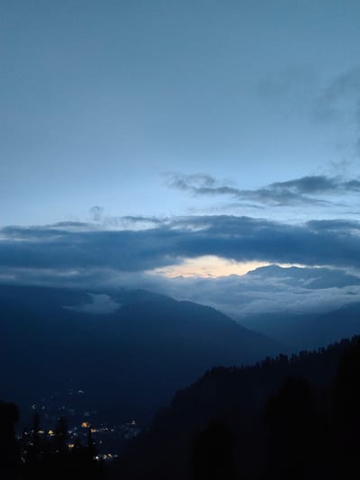The image shows a twilight scene over a mountain range near Aleo, India. The sky is a deep blue with scattered clouds, some of which are dark and ominous, while others are lighter and tinged with the last rays of the sun. In the foreground, silhouetted trees obscure a distant view of a town or village illuminated by artificial lights. The town appears nestled in a valley, with clusters of lights indicating buildings. The mountain slopes are dark and covered in dense vegetation, also silhouetted against the fading light. The overall atmosphere is serene and slightly mysterious, capturing the transition from day to night in a remote, mountainous region.