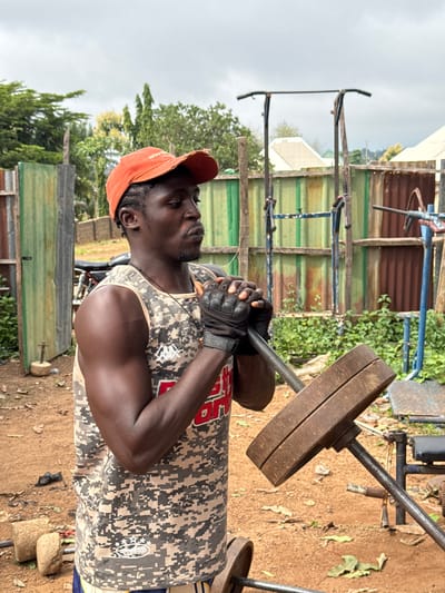 The image shows a man lifting weights outdoors in Akwanga, Nigeria. He is wearing an orange baseball cap, fingerless workout gloves, and a camouflage tank top. The man is focused and appears to be exerting himself as he lifts a barbell with weights. The background includes a makeshift outdoor gym with various pieces of equipment, including a bench and other weights. There are also corrugated metal structures, vegetation, and trees, suggesting a somewhat rural or informal setting. The ground is dirt and scattered with leaves. The lighting indicates it is daytime, and the sky is overcast. There is no discernible text in the image.
