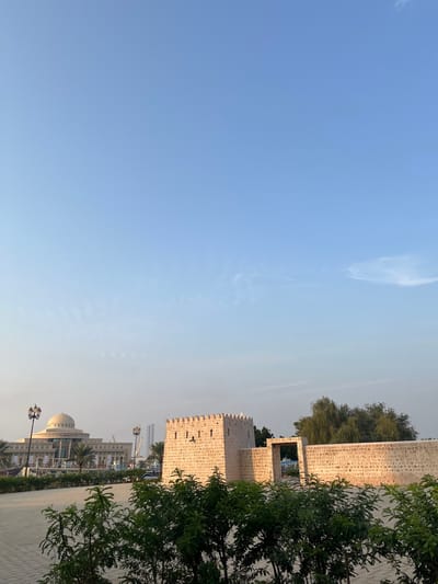 The image displays an outdoor urban landscape in Sharjah, United Arab Emirates, featuring historical architecture juxtaposed with modern buildings under a clear daytime sky. In the foreground, green shrubbery partially obscures a paved ground surface. The midground is dominated by the Al Hisn Sharjah (Sharjah Fort), a light-colored stone structure consisting of a prominent rectangular tower with crenellated battlements and connecting stone walls that incorporate a central arched gateway. A tall, slender tree with green foliage is visible behind the fort's walls to the right. In the background, to the left, stands a large, multi-story contemporary building with a distinctive central dome and columns, identified as the Sharjah Ruler's Office. Ornate, dark-colored streetlights are positioned along a paved area between the fort and the modern building. Faint outlines of other distant structures and construction equipment are discernible further in the background. The sky is clear, light blue with minimal, wispy white clouds. Natural light illuminates the scene, casting a warm hue on the fort. No individuals are present in the image.