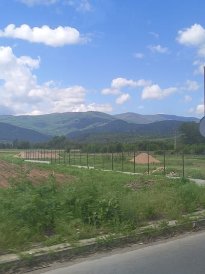 The image shows a landscape in Piperkov Chiflik, Bulgaria. Mountains covered in green vegetation are visible in the background. A blue sky with white clouds is above the mountains. A fence runs along the right side of the image, separating a green field from a road. The road is made of grey concrete and runs along the right edge of the image. The photograph was taken facing the mountains, with the sky to the left.