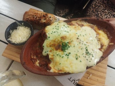 A terracotta dish holds a baked pasta dish, likely lasagna, smothered with a generous layer of melted cheese and sprinkled with herbs. Beside it, a small black bowl contains shredded cheese. A sesame seed-topped bread roll sits near the dish. The meal rests on a wooden board placed on a white, textured surface. The edge of a condiment container appears in the lower-left corner. Part of a leopard-print garment is visible in the background, possibly belonging to a person sitting at the table.