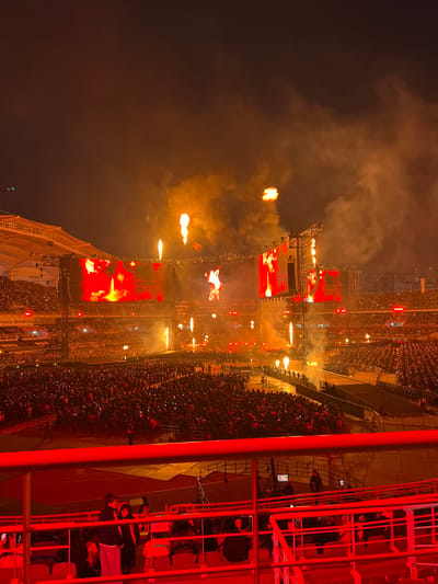 The image depicts a night concert scene at the Goyang Stadium in Goyang-si, South Korea. The stadium is filled with a large crowd of people. A stage is visible, featuring large screens displaying red and orange visuals. There are flames shooting upwards from the stage, and smoke fills the air. The image is taken from an elevated perspective, possibly from seating within the stadium, where a few people are visible in the foreground.