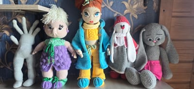 Five crocheted figures are displayed on a wooden shelf against a blue, floral-patterned wallpaper. From left to right, the first figure is a light grey, slender humanoid with large, five-fingered hands. Next to it is a doll with pale skin, white yarn hair, large black eyes with prominent eyelashes, and a purple dress with a green top. To the right of this doll is another doll with red yarn hair, striking blue eyes, wearing a yellow top, blue coat, and yellow pants. Following this is a white and grey crocheted rabbit wearing a red hat and a white scarf. The last figure on the far right is a grey crocheted rabbit with long ears, closed eyes, and a pink nose, wearing a bright pink skirt. The overall scene is an indoor arrangement of handmade toys. The location is the city of Gostagayevskaya, Russia.