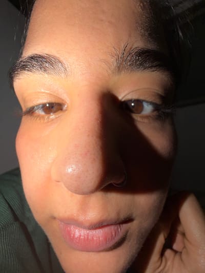 A close-up, angled shot reveals a person's face, featuring dark eyebrows, brown eyes, and a nose piercing. The skin has a slightly uneven tone with visible pores. The lips are pink and slightly parted. Strong shadows cast across the face add depth, obscuring some features. The overall composition highlights the facial features with contrasting light and shadow.