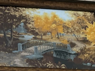 The image is a tapestry depicting an autumn landscape scene with a bridge over a creek. The foliage is rendered in shades of yellow and brown, suggesting the fall season. A stone bridge with arched construction spans a gentle stream. Trees with distinct trunks and branches line the banks of the creek, some with vibrant yellow leaves. The sky above is a pale blue with hints of white clouds. The tapestry is framed by a dark, textured border. There are no people or animals visible in the scene. The artwork is likely a decorative piece, possibly hung in a home or public building. The question about the location in the city of Ponebel, Ukraine, cannot be answered from the image itself, as there are no specific landmarks or identifying features to place it within that city.