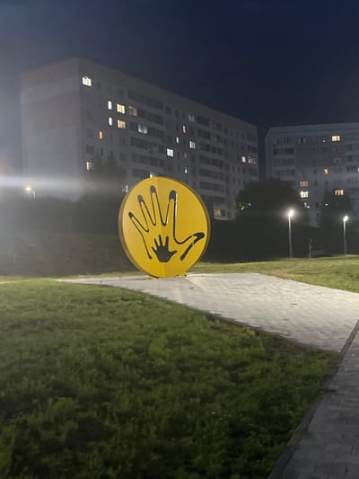 An outdoor nighttime shot captures a yellow, circular sculpture featuring two black handprints, one larger on top of a smaller one. The sculpture is positioned on a paved area adjacent to a grassy lawn. In the background, a multi-story apartment building with illuminated windows is visible, suggesting an urban setting. Two streetlights illuminate the scene, casting a glow on the pathway and surrounding area. The image appears to be taken in the city of Kamskiye Polyani, Russia, a location known for its residential areas and public spaces. The overall atmosphere is serene and somewhat mysterious due to the darkness and artificial lighting.
