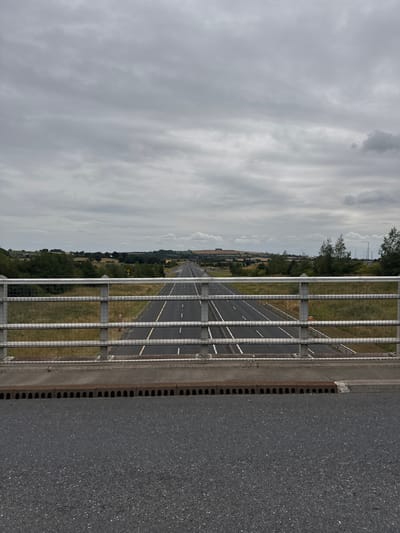 The image depicts a view from a bridge over a motorway in Hacklow, Ireland. The sky is overcast with grey clouds. The motorway stretches into the distance, with multiple lanes marked by white lines. A metal fence with horizontal bars and mesh wire runs along the edge of the bridge, obscuring the view slightly. Beyond the fence, there are grassy verges and trees lining the sides of the road. In the distance, rolling hills and fields are visible, with a few buildings scattered across the landscape. There are no people or vehicles clearly visible in the frame. The foreground shows the asphalt surface of the bridge or an adjacent road, with a metal grate and concrete structure at the edge.