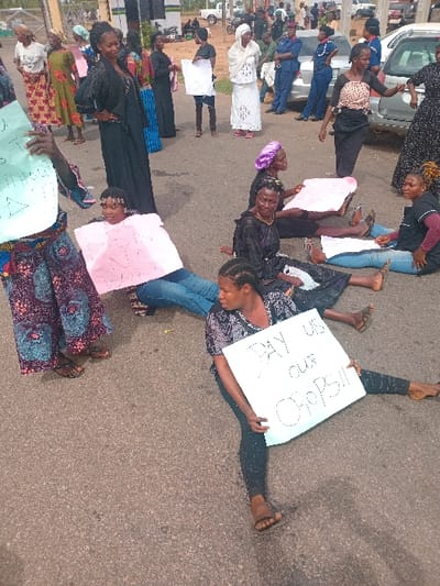 A group of women are protesting in Ungwan Tanko, Nigeria. Several women are seated on the pavement, holding signs with written messages in the local language. One woman in black pants and a black and white shirt is sitting on the ground, holding a sign that reads "PAY us our CROPS!!!". There are two cars parked in the background, and a few men are standing around, observing the protestors. The women appear to be demonstrating for payment for their crops, with some holding signs that read "We need our money" and "We want our money".
