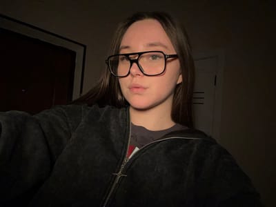 A young adult female is depicted in a dimly lit indoor setting. She has long, dark brown hair and wears black, large-framed glasses. Her facial expression is neutral with slightly parted lips. She is wearing a dark-colored t-shirt underneath an open, textured, dark grey or black zippered hooded jacket. The zipper pull on the jacket is a distinctive silver-colored stylized cross or star. Her right arm is visible, extended towards the left of the frame, suggesting she is holding the device used to capture the image. The background on the left consists of a dark brown door or wall panel with a visible black frame. On the right, a lighter-colored door or wall section features three horizontal parallel lines. The lighting appears to originate from the front, illuminating the subject's face and upper body while the background remains largely in shadow. The location is an indeterminate interior space.